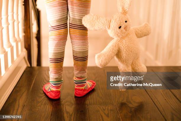 little girl wearing colorful striped leggings & moccasins & holding a soft cuddly stuffed bunny toy at the top of stairs at home - kids feet in home stock-fotos und bilder