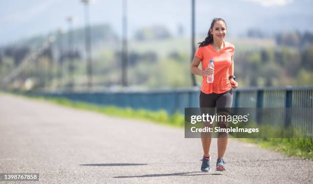 woman in orange shirt and black shorts runs outdoors holding a water bottle on a sunny summer day. - running shorts foto e immagini stock