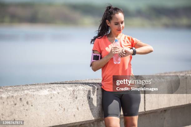 woman is smiling happy with her ruunning performance holding a bottle of water and looking at the watches. - finishing workout stock pictures, royalty-free photos & images