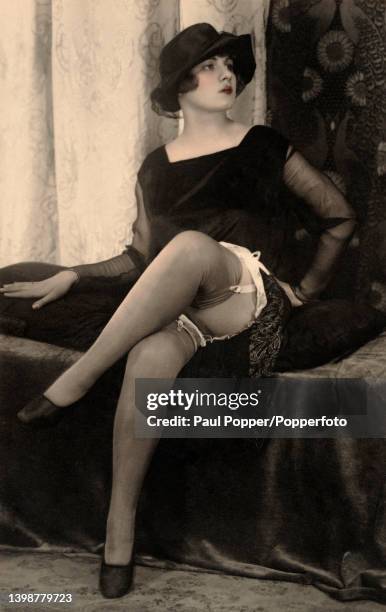 Postcard showing a woman seated on a divan, she wears a square necked black velvet dress with sheer chiffon sleeves and a black cloche hat, her legs...