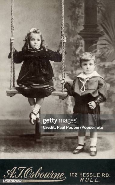 Cabinet card showing two small children in a studio setting, the boy is standing and wears a pleated wool dress with a large sailor style collar, the...