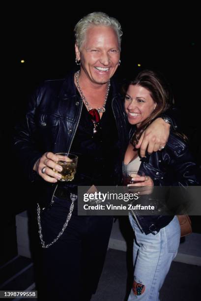 American drummer Matt Sorum, wearing a black leather jacket, a pair sunglasses tucked into the collar of his black shirt, and his arm around a woman,...