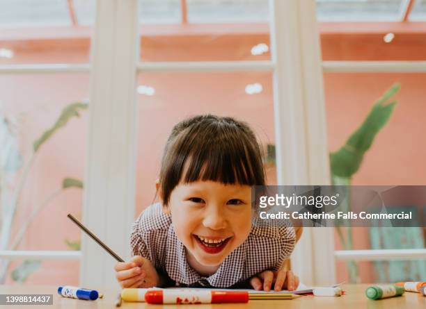 an adorable, happy little girl student leans across a table with a huge smile and looks directly at the camera. she grips a pencil. - escola infantil imagens e fotografias de stock