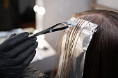 Close-up of a strand of female hair on a special sheet of foil, the master applies hair dye with a brush. The process of highlighting by a professional master in a beauty salon.