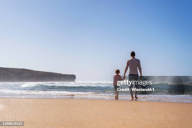 father and daughter walking towards sea on sunny day - children por stock-fotos und bilder