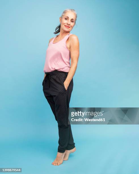 smiling woman with hands in pockets standing against blue background - man portrait full body 50's stock-fotos und bilder