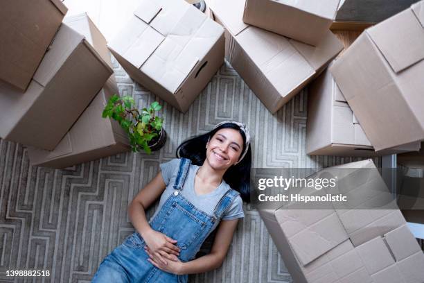 beautiful young woman taking a break from unpacking boxes lying down on the floor facing camera smiling with a toothy smile - moments daily life from above imagens e fotografias de stock