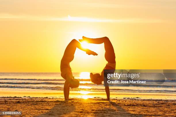 two women doing yoga on the beach at sunrise in the  handstand posture - handstand beach stock pictures, royalty-free photos & images