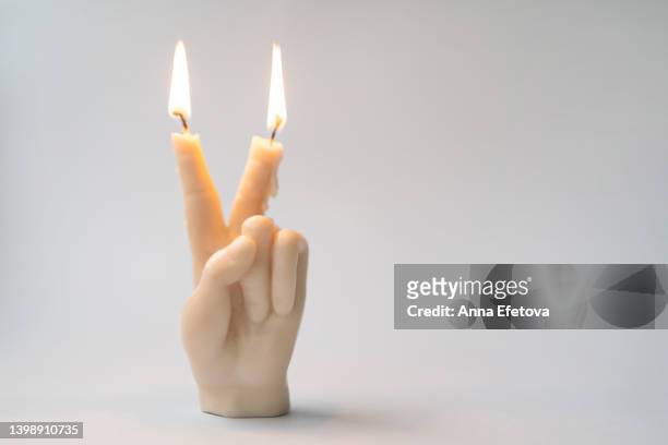 white burning soy or wax candle in shape of a hand gesturing v sign on gray background. concept of peace and tolerance - candle white background stock-fotos und bilder