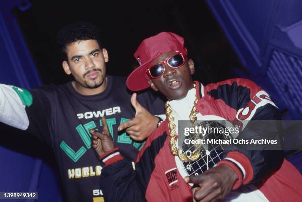 American rapper Flavor Flav, wearing a red baseball cap and sunglasses with a thick gold chain and a clock hanging around his neck, and a man,...