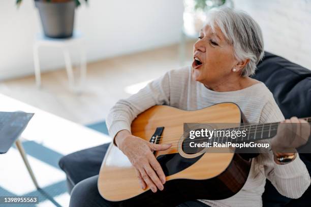 senior woman in casual clothes is smiling while playing guitar at home - woman playing guitar stock pictures, royalty-free photos & images