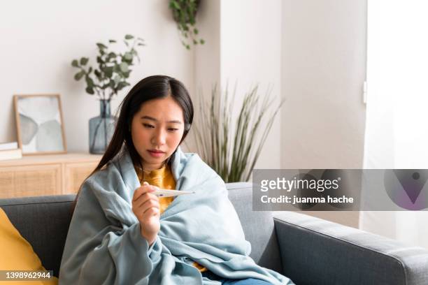 sick woman at home covered with a blanket while using thermometer to take her temperature. - body temperature stock pictures, royalty-free photos & images