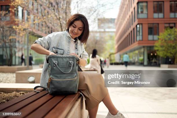 young woman opening her backpack while sitting in park - bolso abierto fotografías e imágenes de stock