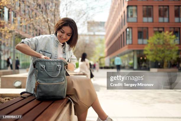 young woman opening her backpack while sitting in park - borsetta beige foto e immagini stock