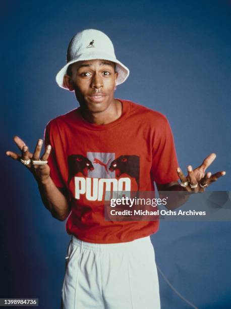 American rapper MC Shan , wearing a red Puma t-shirt with a white Kangol bucket hat and white trousers, in a studio portrait against a blue...