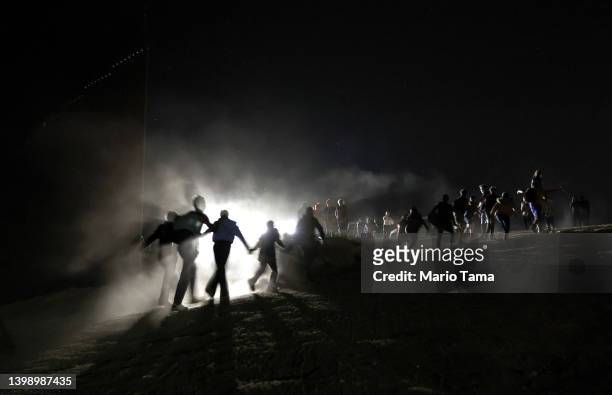 Immigrants cross through a gap in the U.S.-Mexico border barrier as others watch from above before being processed by the U.S. Border Patrol on May...