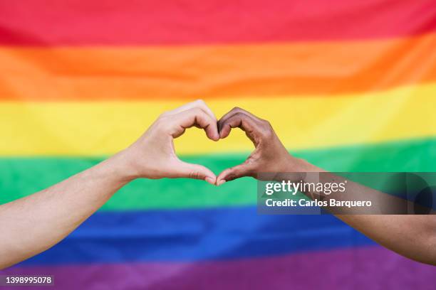 photo of two hands doing a heart and a lgtbia+ flag in the background. - gay pride flag foto e immagini stock