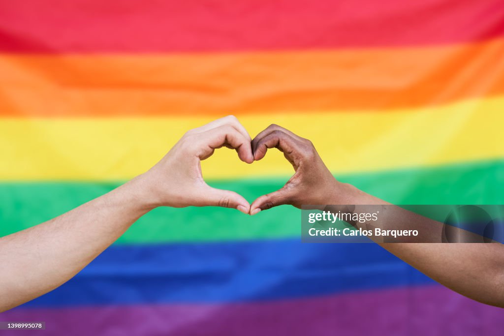 Photo of two hands doing a heart and a Lgtbia+ flag in the background.