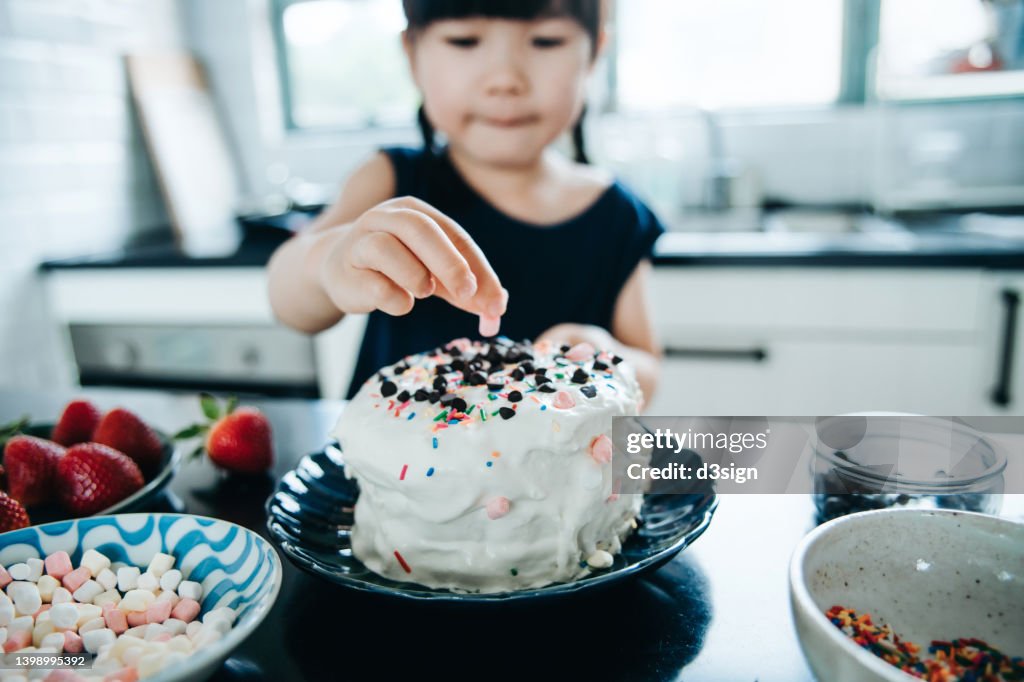 Little Asian girl baking cake at home, decorating cake with fresh strawberries, marshmallow, chocolates and sugar sprinkles at kitchen counter. DIY. Baking at home. Birthday and celebration concept