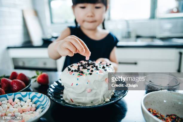 little asian girl baking cake at home, decorating cake with fresh strawberries, marshmallow, chocolates and sugar sprinkles at kitchen counter. diy. baking at home. birthday and celebration concept - cake decoration stock pictures, royalty-free photos & images