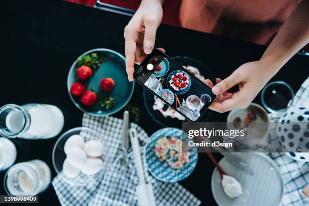 high angle shot of young woman baking cake at home, taking photos of her freshly homemade cake with smartphone. with assorted baking supplies on kitchen counter. baking at home. diy. birthday and celebration concept - fotohandy stock-fotos und bilder