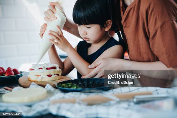 asian mother and little daughter baking a cake and decorating with strawberry and whipped cream together in the kitchen. family baking at home. diy. birthday and celebration concept - cremetorte stock-fotos und bilder