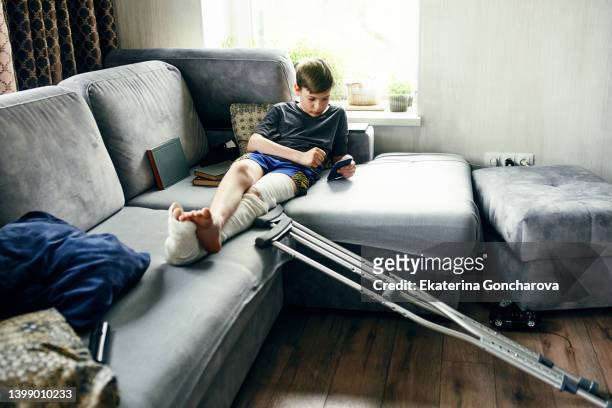 a boy with a broken leg and crutches is sitting on a sofa and using mobile phone - broken cell phone stock-fotos und bilder