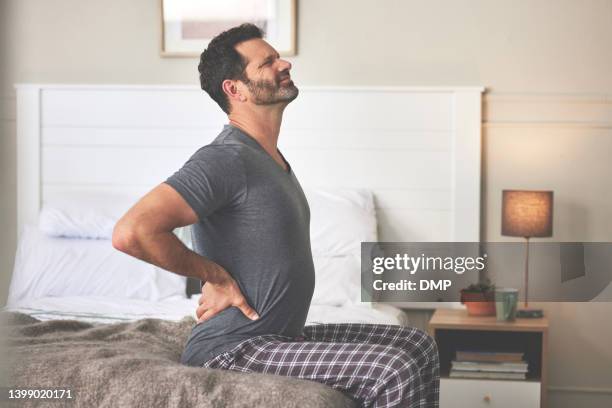 a man feeling back pain in bed at home. a mature guy looking unhappy, stressed and feeling sick in his bedroom during the morning - stijf stockfoto's en -beelden