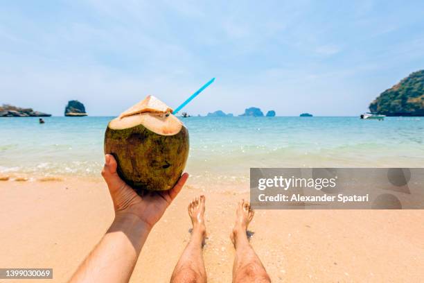 man drinking fresh coconut milk while relaxing by the ocean at the beach, personal perspective view - beautiful male feet stock pictures, royalty-free photos & images