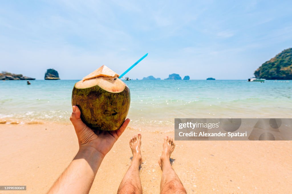 Man drinking fresh coconut milk while relaxing by the ocean at the beach, personal perspective view