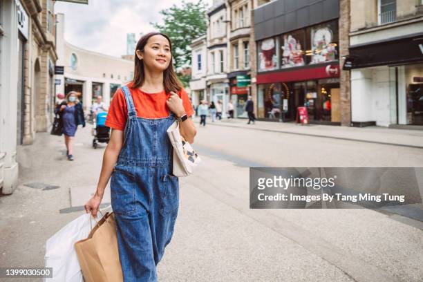 young cheerful asian woman carrying shopping bags while walking in the high street in downtown district - uk high street shops stock pictures, royalty-free photos & images
