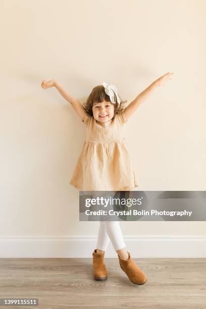 a cute 3-year-old toddler girl standing and posing while wearing a yellow plaid shirt, white pants, brown boots & a white hair bow - beige shirt stock pictures, royalty-free photos & images