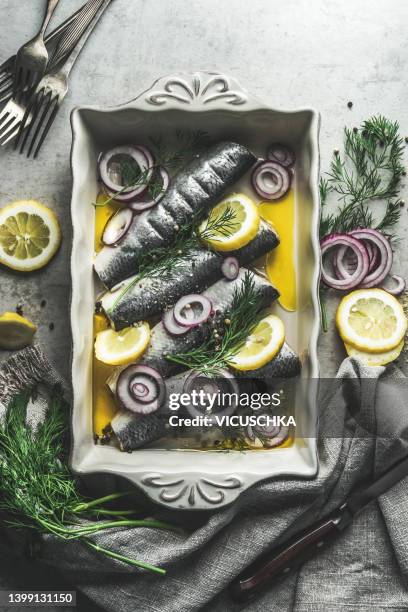 raw fishes in casserole with dill, lemon and red onion at grey concrete kitchen table with forks. - arenque fotografías e imágenes de stock