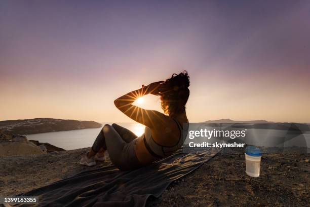 female athlete doing sit-ups on a hill above the sea at sunset. - cyclades islands stock pictures, royalty-free photos & images