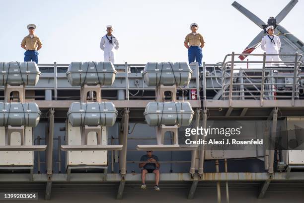 Marines and Navy sailors from the USS Bataan stand on the flight deck above another man sitting off the edge during their arrival for Fleet Week 2022...