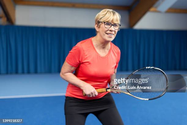 happy senior woman playing tennis on an indoor court - atuendo de tenis fotografías e imágenes de stock