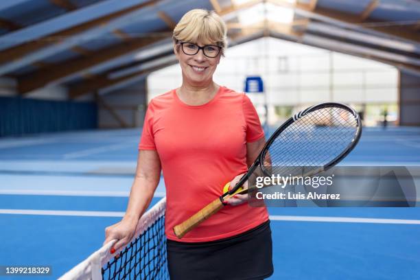 healthy senior woman with racket standing by net on tennis court - woman short blond hair stock pictures, royalty-free photos & images