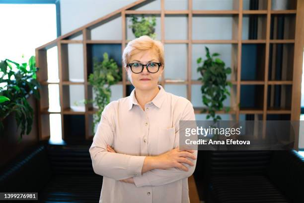 smiling senior blond haired woman with glasses stands in office minimalist interior. she is wearing light colored shirt - russian business woman stock-fotos und bilder
