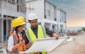 Caucasian males and Asian female builders, architector and engineers with draft plan of building and laptop computer talking on constructing site
