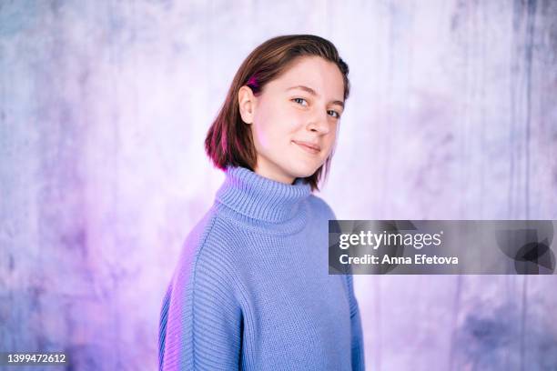 beautiful young woman smiling against gray wall background with violet neon lighting. she has short hair and gray eyes. she is wearing a purple sweater. concept of natural beauty - lavender color fotografías e imágenes de stock