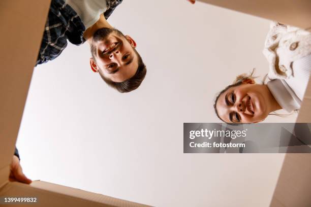 low angle view of couple looking at camera that is inside the cardboard box they are unpacking - inside of box stock pictures, royalty-free photos & images