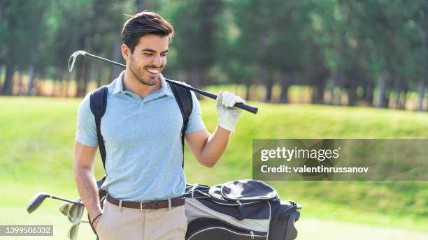 joven jugador de golf sonriente sosteniendo un palo de golf en su hombro, de pie junto a sus palos en una bolsa en un campo de golf. - bolsa de golf fotografías e imágenes de stock