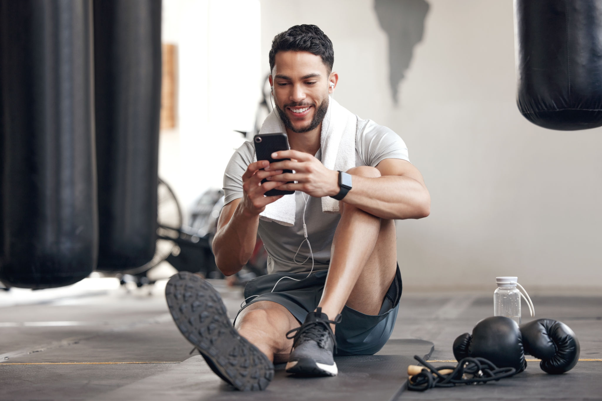 One fit young hispanic man listening to music with earphones from a cellphone while taking a break from exercise in a gym. Happy guy texting, making video call and using fitness apps online while browsing social media and watching workout tutorials One fit young hispanic man listening to music with earphones from a cellphone while taking a break from exercise in a gym. Happy guy texting, making video call and using fitness apps online while browsing social media and watching workout tutorials