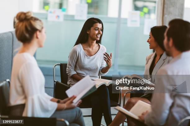 joven mujer de etnia india liderando una discusión mientras repasa libros en reunión en un espacio interior moderno - club de lectura fotografías e imágenes de stock