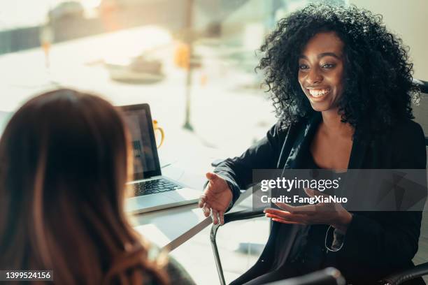 black woman in casual work attire talking to business colleague while sitting in chair in modern office with laptop on desk table - recruiter stock pictures, royalty-free photos & images