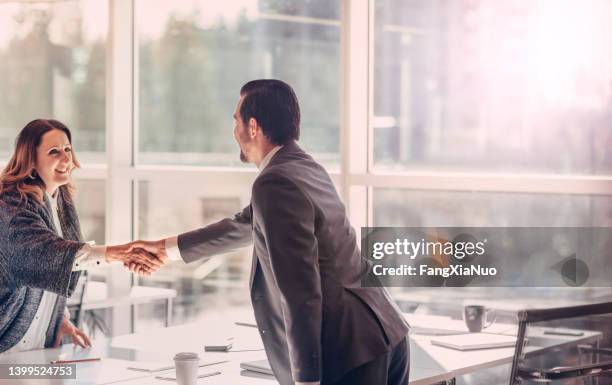 man and woman standing shaking hands across a table in agreement job interview success inside a modern business office - initiative stock pictures, royalty-free photos & images