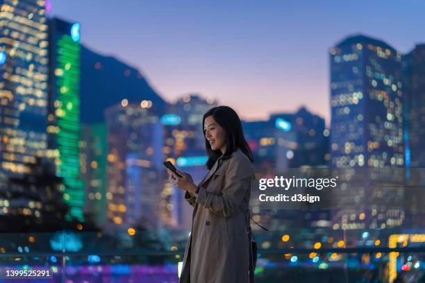 young asian businesswoman using smartphone in downtown city street. confident female executive using smartphone in prosperous financial district, against illuminated and multi-coloured corporate skyscrapers at night. business on the go. female leadership - central asia stock-fotos und bilder