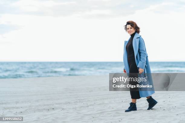 lonely woman strolling along seaside, on sandy beach, smiling at camera. copy space - clumsy walker stock pictures, royalty-free photos & images