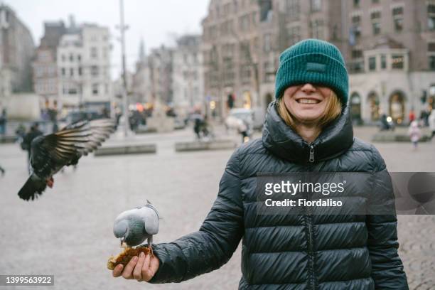 laughing cheerful woman in the main square of the old city feeding a flock of doves with rice - amsterdam autumn stock pictures, royalty-free photos & images