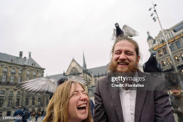 laughing cheerful man and woman in the main square of the old city feeding a flock of pigeons with rice - mature couple winter outdoors stock pictures, royalty-free photos & images